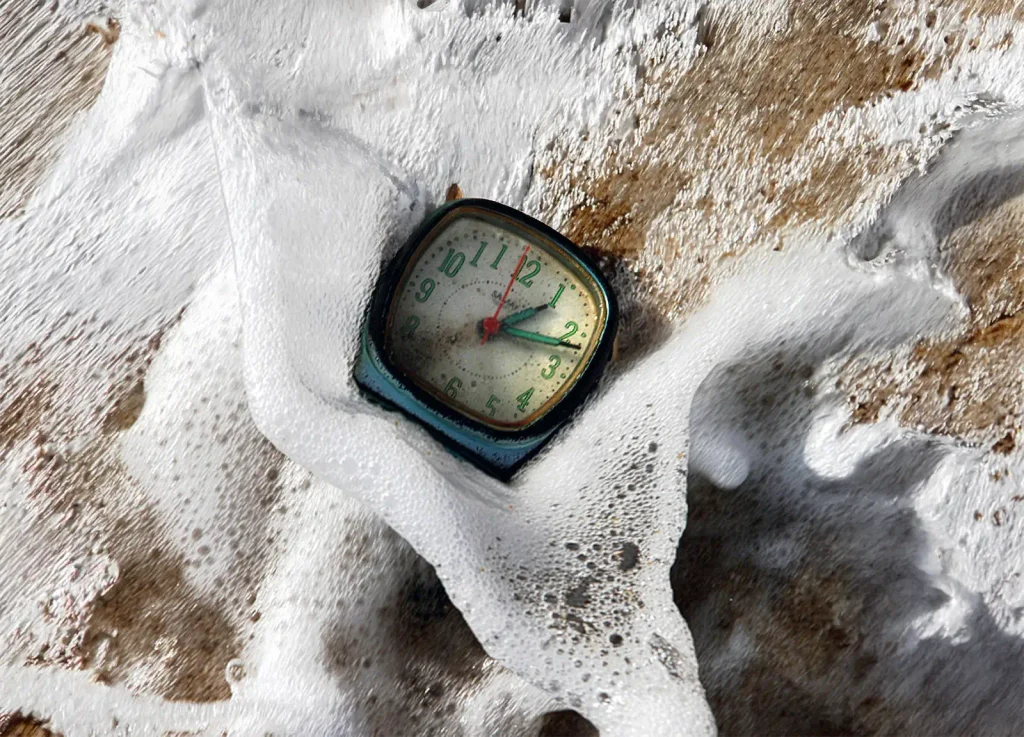 Old analog clock resting in white sea foam on a sandy beach